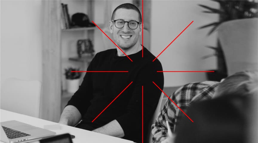 Immagine in bianco e nero di un uomo sorridente con il computer. Un elemento grafico a linee rosse dal centro.